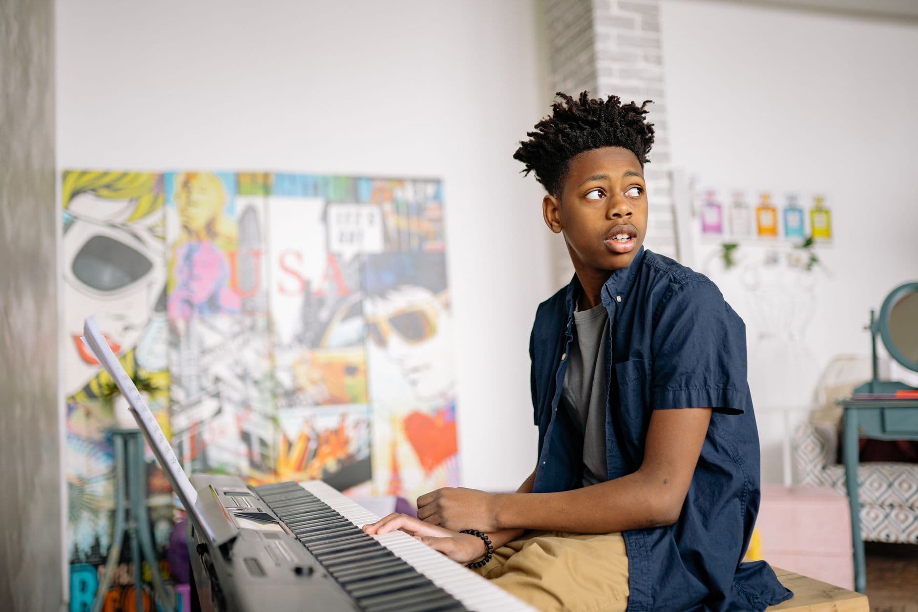 A teenager with afro hair playing the piano in a creatively decorated room, looking away.