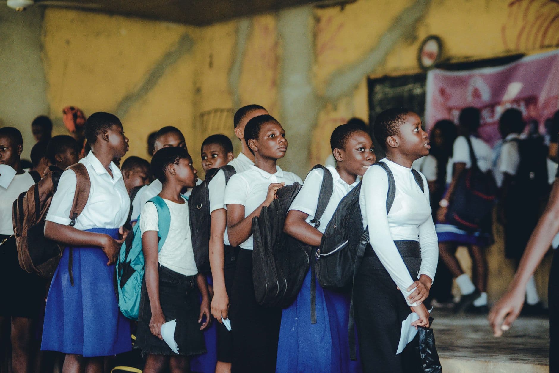A group of schoolchildren in uniform lining up indoors, carrying backpacks.
